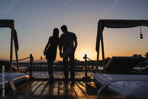 Young romantic couple in love watching sunset together on the beach. Traveling, summer holidays. Sunset beach. People silhouette from behind, standing and enjoying view of the sea.