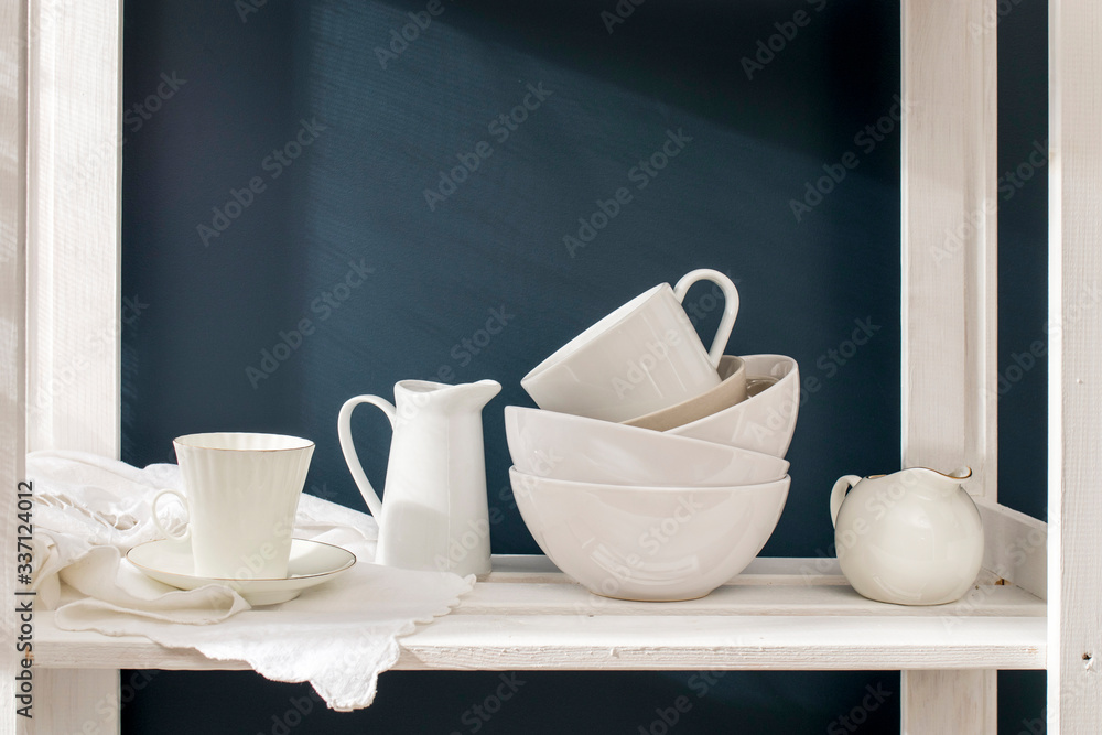 A a set of white dishes: porcelain milk jug, coffee cup, bowls on a white rack on a dark blue background