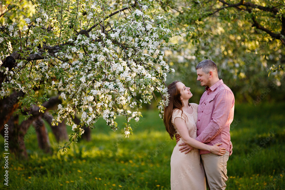 Young man and woman couple in a blooming apple garden. Tender holding each other. Spring lovestory. Brown-haired girl with long hairs and man in pink shirt. Young family. Couple romantic spring date