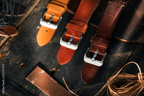 Handmade brown tone watch straps with steel buckle laying on wooden rustic surface next to leather cuttings and leather thread. Different leather colour tones.