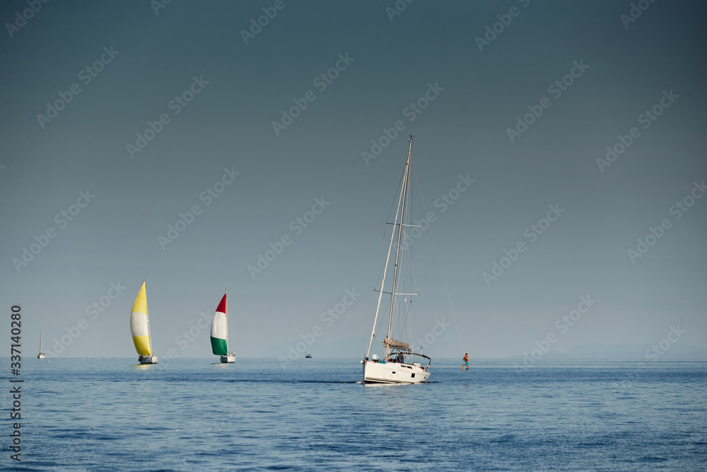 Obraz premium Members of team have a good time after completion of race and dive into water from a rope on a mast, The race of sailboats, Intense competition, bright colors, island with lighthouse are on background