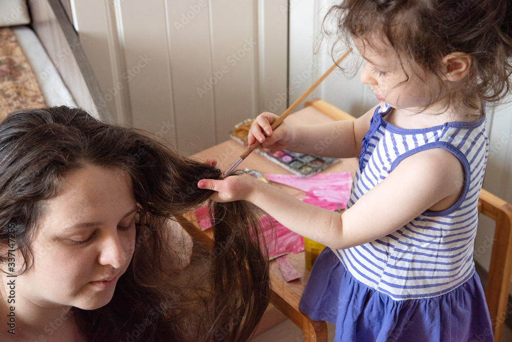 Fototapeta premium what to do in quarantine. child paints her mother s long hair with paints. Children and parents during self isolation.Mom sits on the floor, a little girl dyes her hair with watercolors. Happy family