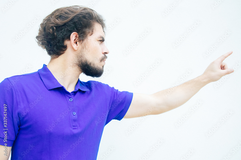 Angry displeased guy pointing finger away. Handsome bearded young man in blue casual t-shirt posing isolated over white background. Get out or fired concept Angry displeased guy pointing finger away. Handsome bearded young man in blue casual t-shirt posing isolated over white background. Get out or fired concept