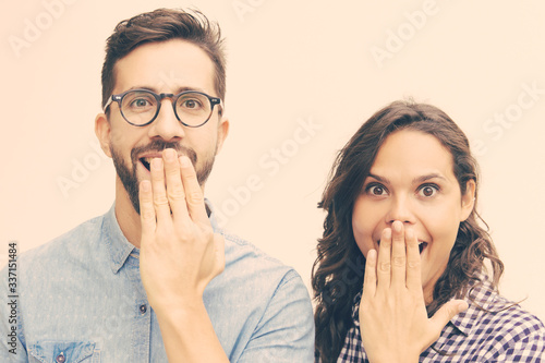 Surprised happy couple staring at camera and covering open mouths with hands. Young woman in casual and man in glasses standing isolated over white background. Great news concept