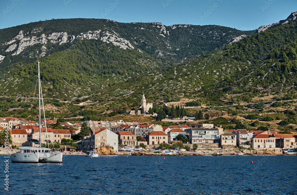 Seascape of city Komiza - the one of numerous port towns in Croatia, Catamaran in the foreground, orange roofs of houses, a cathedral St.Nicholas, Picturesque slopes of mountains