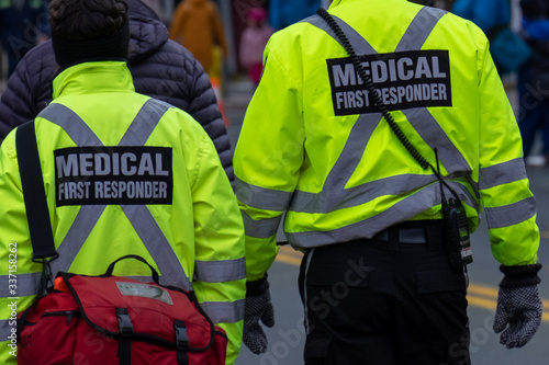 Two medical first responders walking in a street among a crowd of people.One of the EMTs is carrying a red first aid bag.The emergency personnel have bright yellow reflective jackets with grey stripes photo