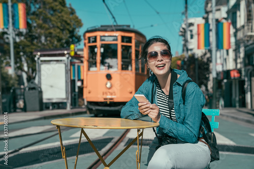 cheerful lady traveler with camera sitting at outdoor cafe table using mobile phone watching funny movie. tourist relax on road with cable car driving in back. rainbow flag hanging on castro street