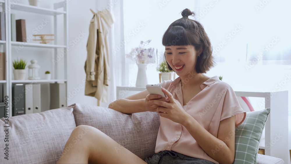 Charming pretty girl sitting on sofa in bright living room at home. young happy asian japanese woman doing online shopping through 3G internet having smart phone in hands relax on couch indoors. Charming pretty girl sitting on sofa in bright living room at home. young happy asian japanese woman doing online shopping through 3G internet having smart phone in hands relax on couch indoors.