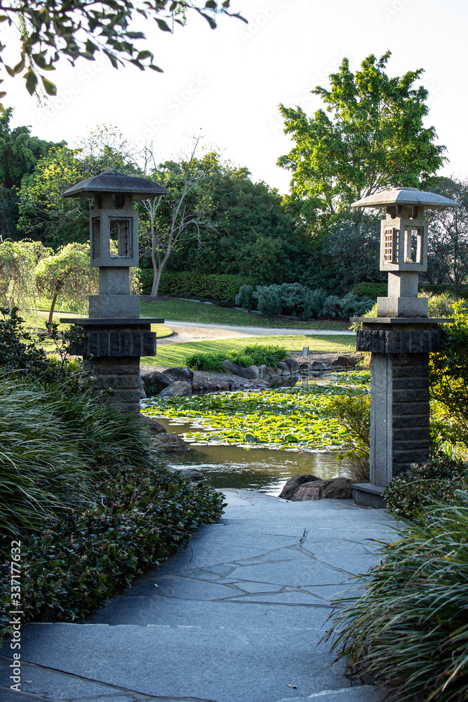 Winding garden path with Chinese style lanterns on pillars leading to water lilies floating on pond with trees in background