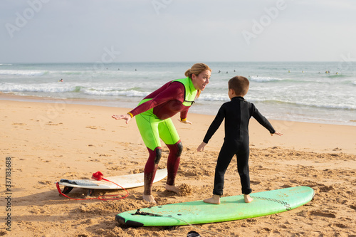 Excited mother with open mouth playing with son. Woman standing on sand beach near surfboards. Lady and boy spreading arms. Sea waves with foam on background. Vacation, surfing and summer concept