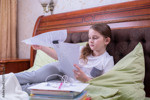 A schoolgirl on bed at home reads text on sheet of paper, undergoes distance learning.