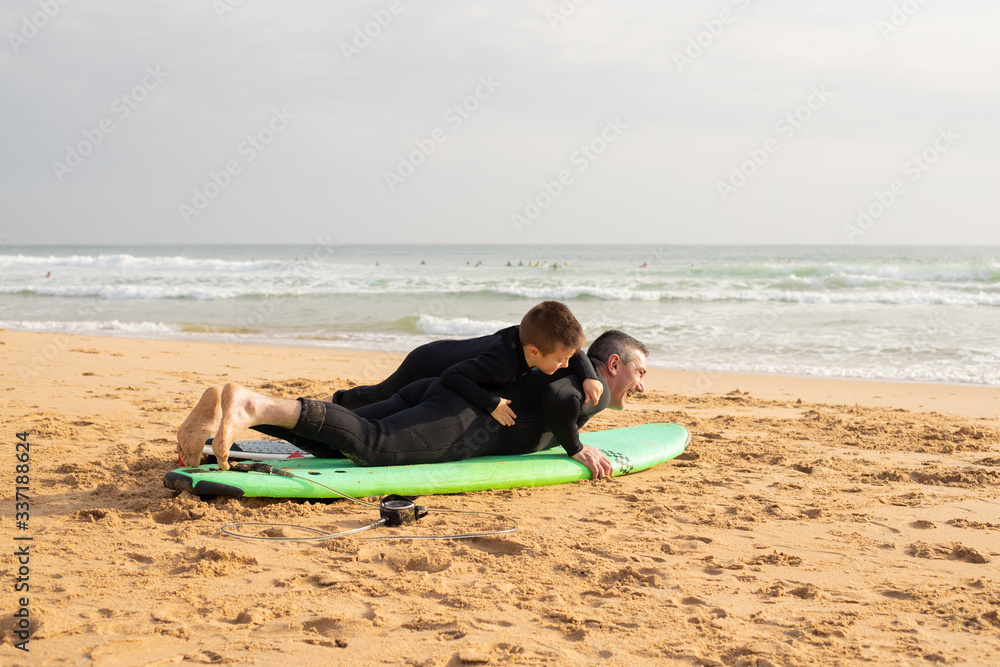 Father laying on surfboard and playing with son. Smiling middle-aged man and little boy in full body swimsuits. Sea waves with foam on background. Vacation, surfing and summer concept