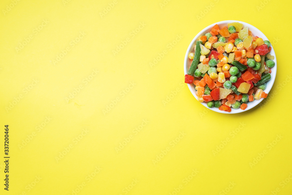 Chopped frozen vegetables in a plate on a yellow background. Corn peas pepper carrots. copy space.