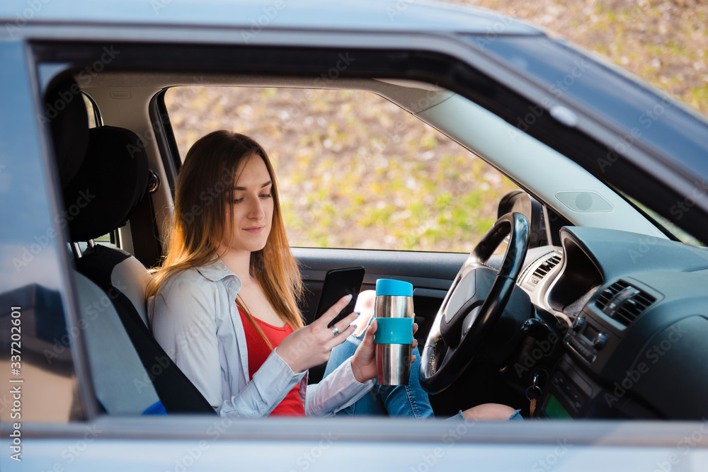 Young woman sitting at the wheel and using a mobile phone in the afternoon. In his other hand he holds a thermo cup with coffee
