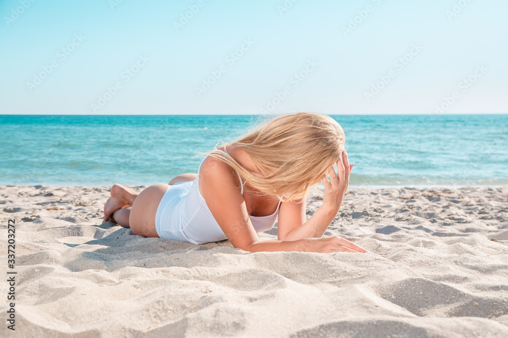 Girl with blond hair basking in sand by sea. A woman sunbathes on summer beach.