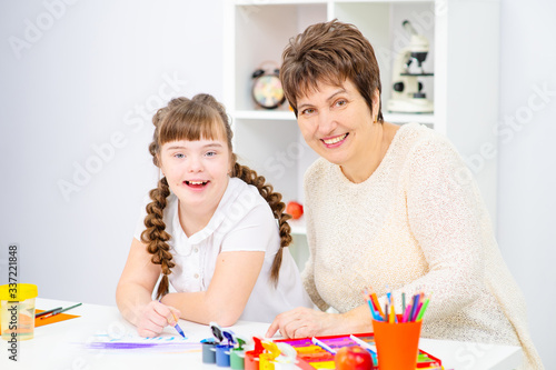 The girl and mother sitting at the table draw a picture at home. A girl with down syndrome at home with her mother is engaged in creativity