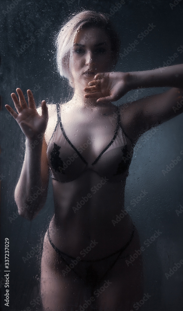 Girl blonde in underwear behind wet glass against a dark background.