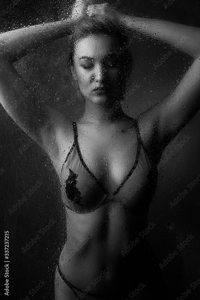 Naklejka premium Girl blonde in underwear behind wet glass against a dark background.