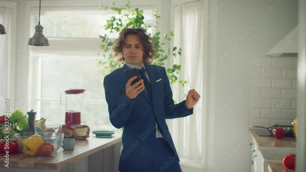 Happy Young Man with Long Hair Dancing at Home while Wearing Blue Business Suit. He is Listening to Music on a Mobile. Energetic Man Using Smartphone in a Kitchen with Healthy Vegetables.