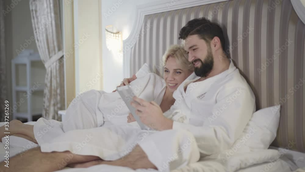 Side view of happy young man using tablet and hugging smiling blond woman in bed. Joyful Caucasian couple resting in bedroom and surfing social media. Bonding, love, leisure, lifestyle. Side view of happy young man using tablet and hugging smiling blond woman in bed. Joyful Caucasian couple resting in bedroom and surfing social media. Bonding, love, leisure, lifestyle.