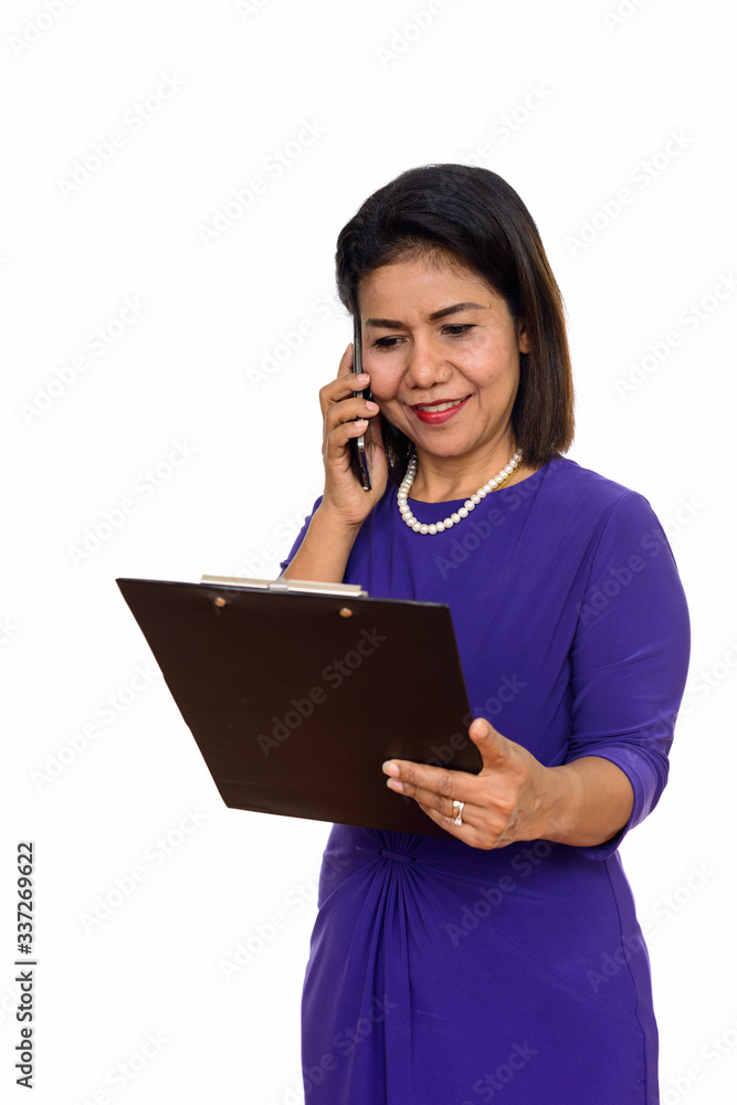 Studio shot of happy mature Asian woman holding clipboard while talking on mobile phone Studio shot of happy mature Asian woman holding clipboard while talking on mobile phone