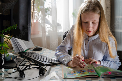 Teenager girl with long blond hair learns via desktop computer from home room.  Girl look at and read student book. Distance electronic learning online education.  

