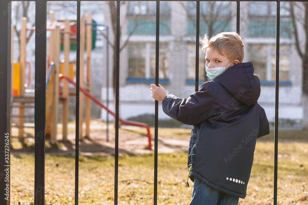 Obraz premium A boy in a medical mask stands next to a metal fence. A child yearns for playing on the Playground during quarantine