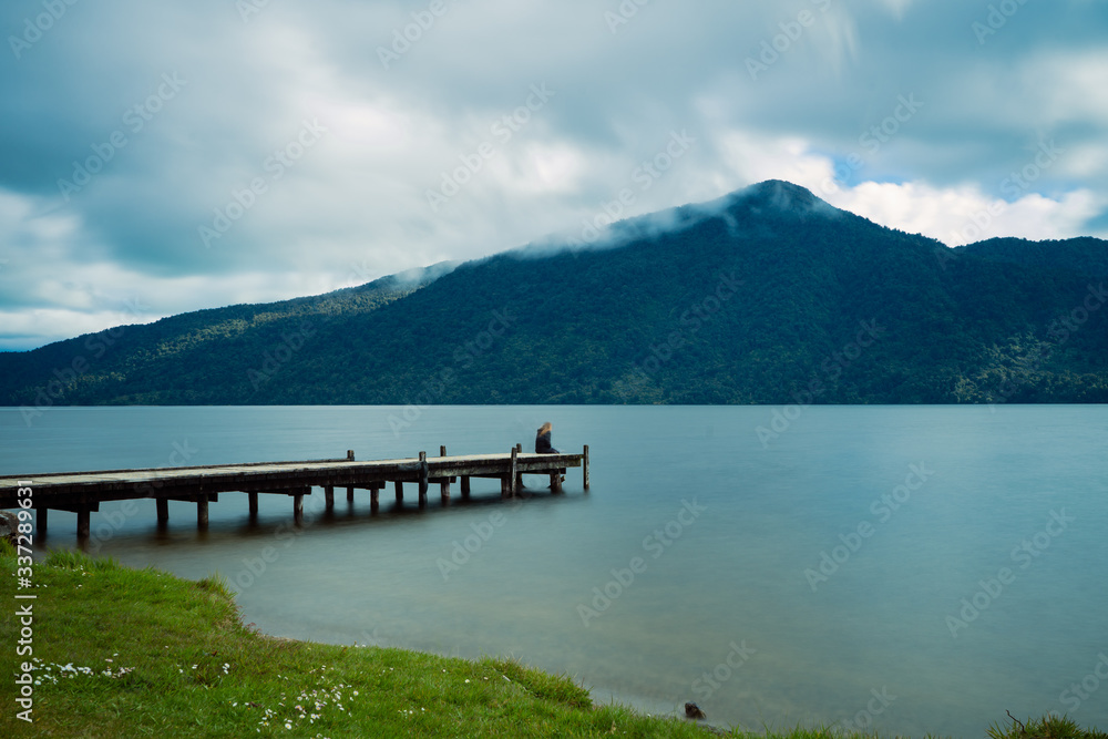 Kaniere Lake, New Zealand, October 7, 2019: Beautiful view of a European woman sitting at the end of the pier admiring the scenery
