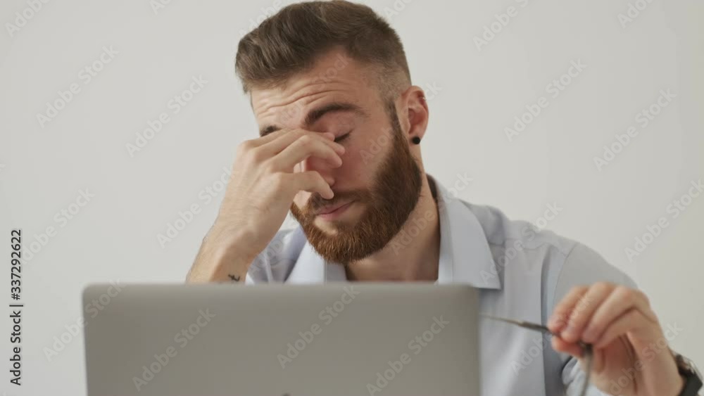 A tired young bearded man put off his eyeglasses while working with his laptop computer sitting at the table over white wall A tired young bearded man put off his eyeglasses while working with his laptop computer sitting at the table over white wall
