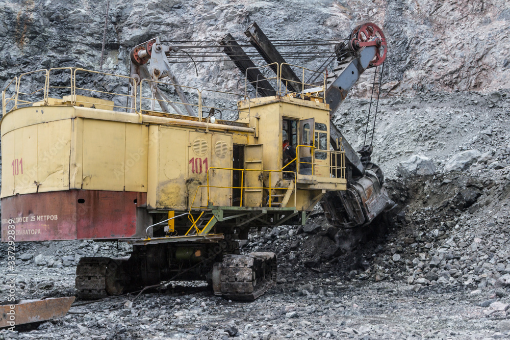 A daytime view of the iron ore mining quarry. In sunny weather. A heavy-duty heavy excavator loads ore onto a dump truck for transportation on an open iron ore mining career. A daytime view of the iron ore mining quarry. In sunny weather. A heavy-duty heavy excavator loads ore onto a dump truck for transportation on an open iron ore mining career.