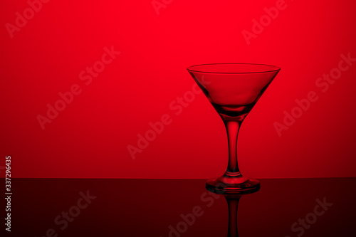 Glass goblet without wine on a thin leg stands on a mirror surface. Back light. Red background. Copy space.