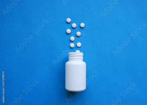 A pile of pills on a blue background. White pills fall out from a white jar on a blue background.