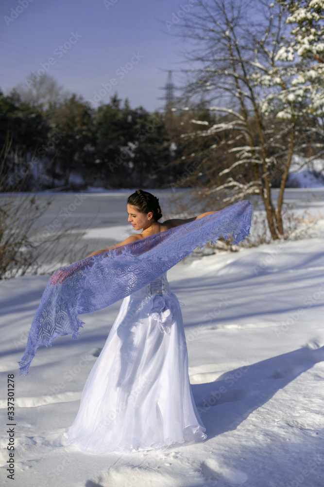 vertical portrait in the winter forest of a slender brunette in a long white dress and a beautiful openwork lilac shawl