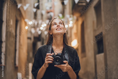 Outdoor summer smiling lifestyle portrait of pretty young woman having fun taking pictures in the city during her travel. She is using professional photo camera and looking around beautiful streets