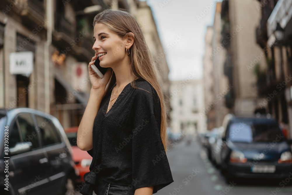 Fototapeta premium Young woman looks startled receiving some information by the call on her phone. She listen to the audio message and became happy hearing the voice of her friend. Girl is crossing the road.
