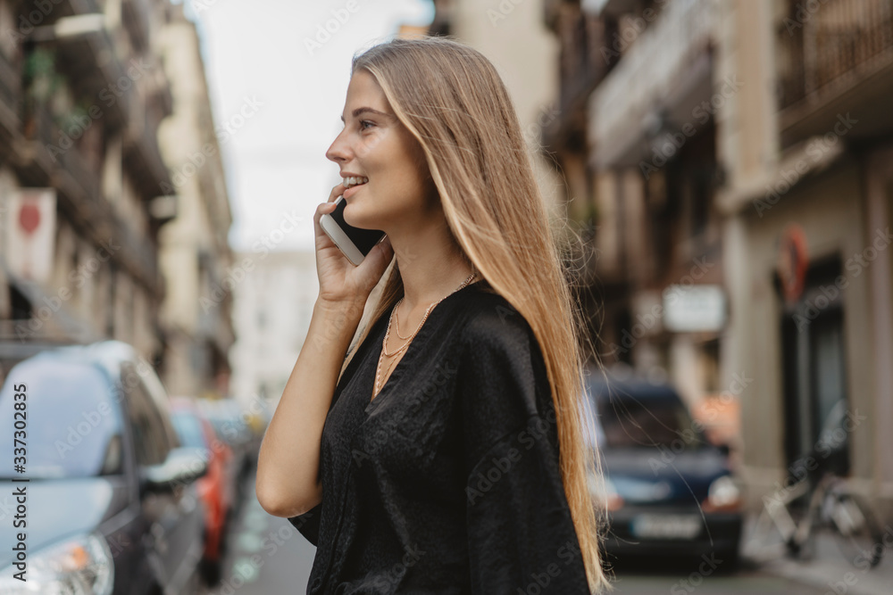 Fototapeta premium Girl crosses the road with cars and speak with her friend by the cellphone, she smiles and looks happy. Woman is using her gadget to communicate with her friends.