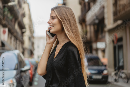 Girl crosses the road with cars and speak with her friend by the cellphone, she smiles and looks happy. Woman is using her gadget to communicate with her friends. © makarovada