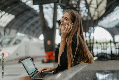 Girl is holding her smartphone and accepting a call. Young blonde woman is sitting on the stone bench at the railway station and using her grey laptop for chatting with her friends.