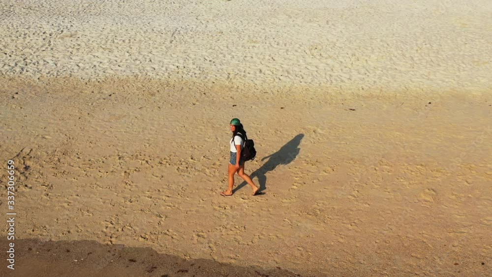 Girl with backpack walking on sand of exotic beach alongside sea on a sunny morning making long shadow in Bahamas Girl with backpack walking on sand of exotic beach alongside sea on a sunny morning making long shadow in Bahamas