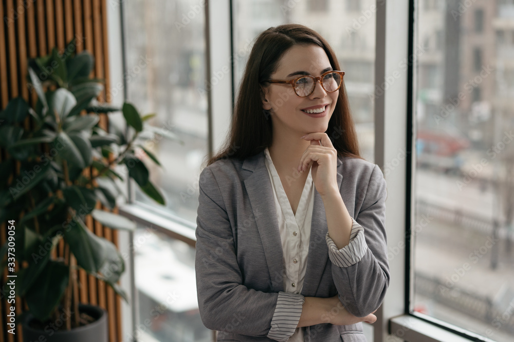 Fototapeta premium Young confident manager standing in office near window, smiling. Portrait of successful business woman in formal clothes and stylish eyeglasses planning start up at workplace