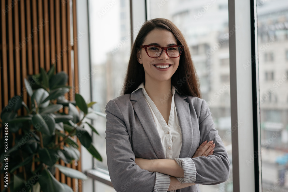 Portrait of young confident business woman with arms crossed standing in office. Successful manager in formal clothes and stylish eyeglasses looking at camera and smiling Portrait of young confident business woman with arms crossed standing in office. Successful manager in formal clothes and stylish eyeglasses looking at camera and smiling
