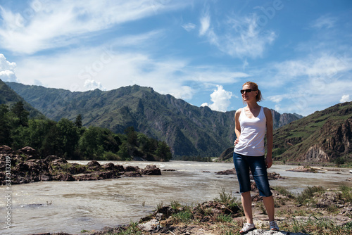 Happy female tourist in a white t-shirt and denim shorts stands on bank of mountain river on sunny day. Cheerful young girl travels in the mountains and admires the landscape.