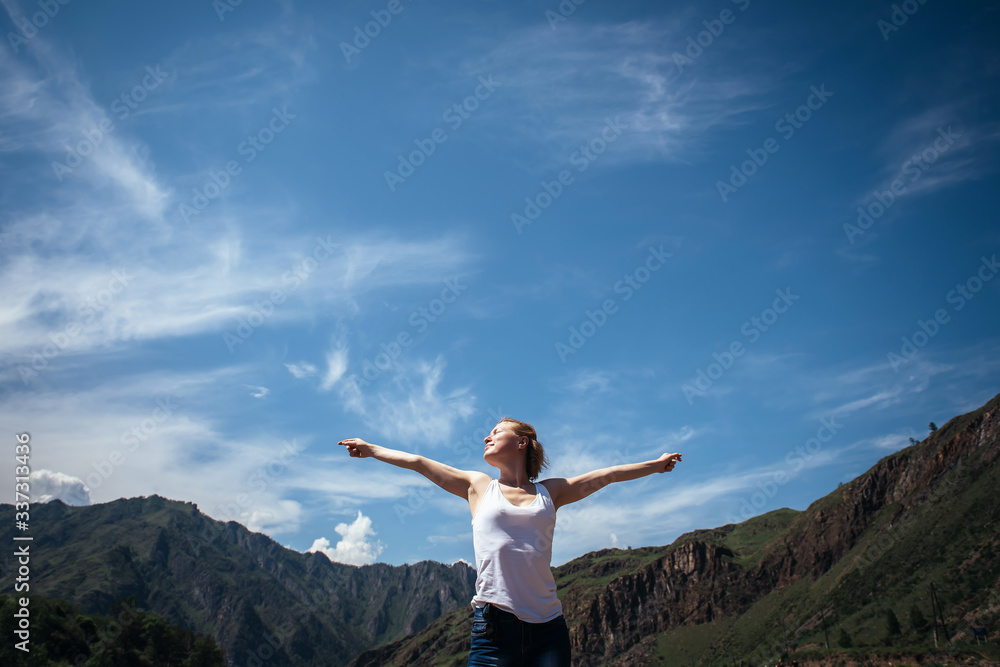 Happy female traveller in a white t-shirt against the beautiful mountains and blue sky on sunny day. Cheerful young girl enjoying a trip in the Altai mountains. Happy female traveller in a white t-shirt against the beautiful mountains and blue sky on sunny day. Cheerful young girl enjoying a trip in the Altai mountains.