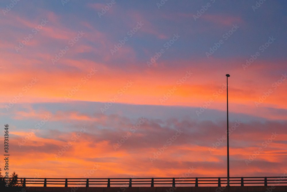 Silueta de farola y valla de carretera sobre cielo naranja con nubes al sol del atardecer