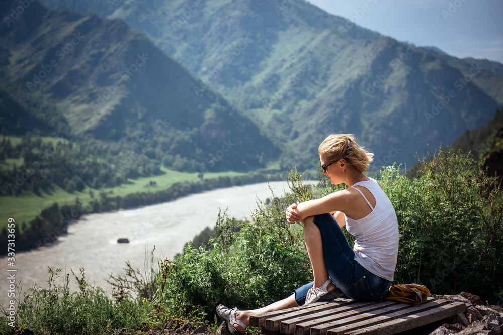 Young woman in white t-shirt and shorts sits on top of hill and admires the beautiful view of the mountains and river on snny summer day. Pretty blonde resting while hiking in the mountains.