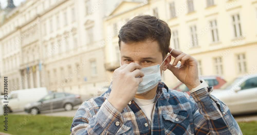 Close up of young handsome Caucasian man wearing white medical mask as protection from coronavirus and looking at camera. Outdoors. Portrait of guy taking on protective antivirus mask. Close up of young handsome Caucasian man wearing white medical mask as protection from coronavirus and looking at camera. Outdoors. Portrait of guy taking on protective antivirus mask.