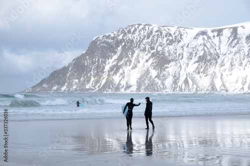 Surfing in cold ocean in Lofoten islands. Famous destination for surfers all around the world. Surfing between snowy mountains and arctic beaches. Surfer's couple is going in to the ocean.