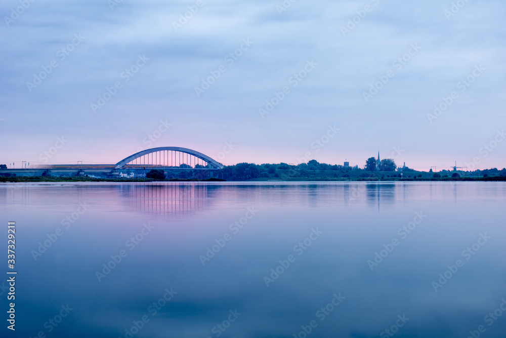 Fototapeta premium Long exposure of a view on Culemborg The Netherlands over the river Lek with railway bridge and train crossing blue sky and water pink sunrise