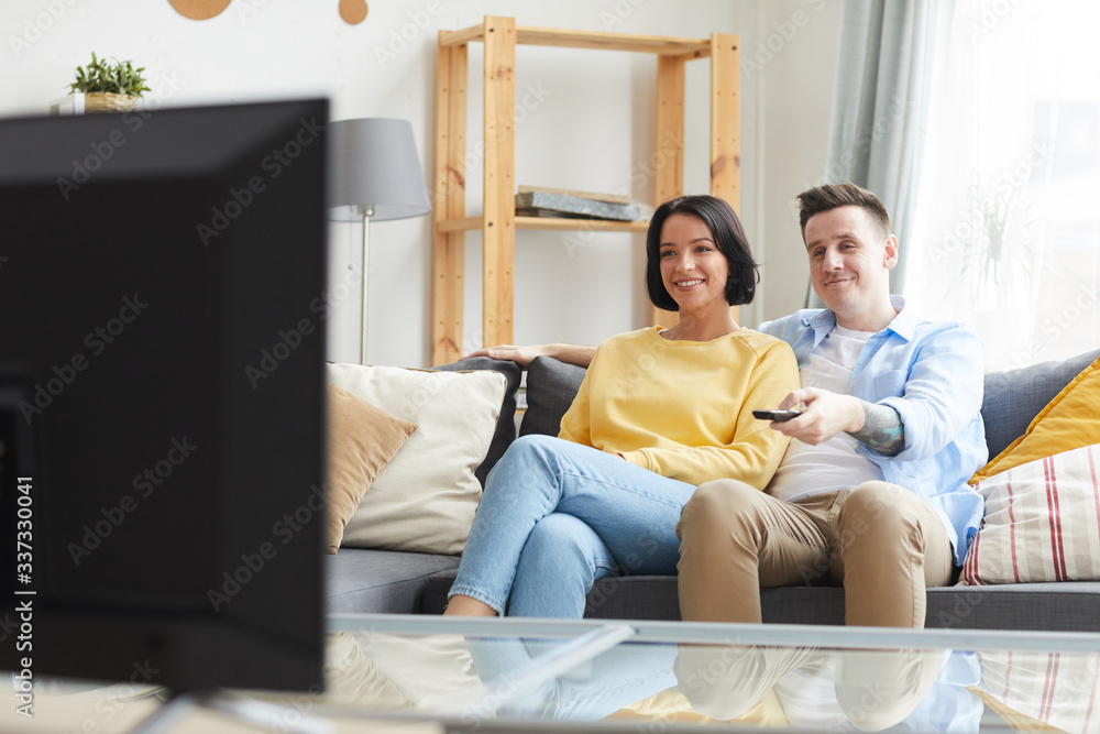 Young smiling couple sitting on sofa and watching TV in the living room at home Young smiling couple sitting on sofa and watching TV in the living room at home