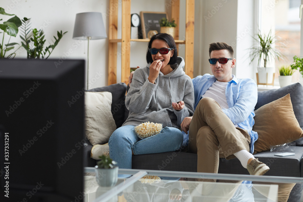 Obraz premium Happy young couple wearing 3D glasses sitting on sofa and eating popcorn while watching the movie on TV